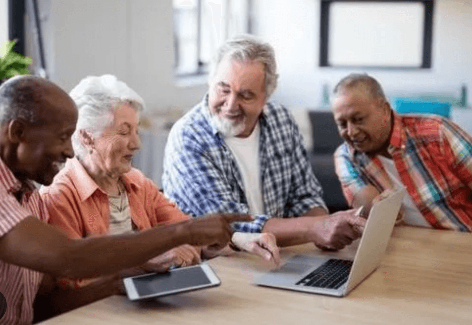 Four older adults gathered around a laptop and tablet at a table, laughing as one of them points at the screen